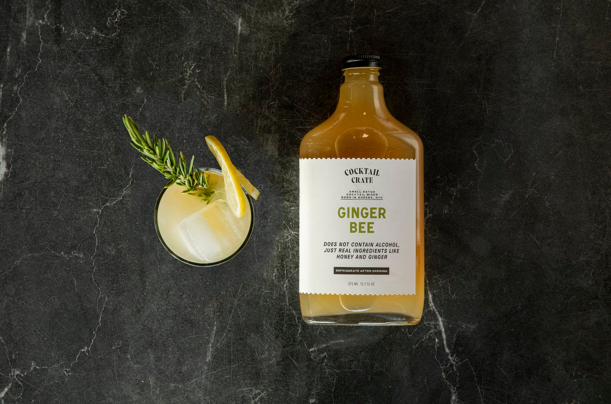 Cocktail Crate Ginger Bee mixer bottle beside a Ginger Bee whiskey cocktail with rosemary and a lemon slice on dark marble