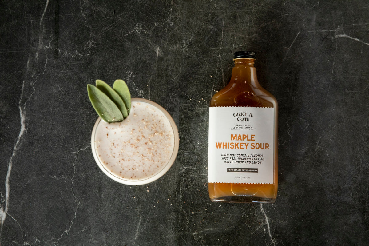 Cocktail Crate Maple Whiskey Sour mixer bottle beside a Maple Whiskey Sour cocktail with a sage garnish on dark marble