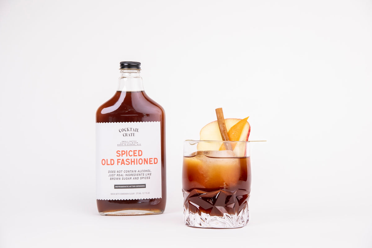 Cocktail Crate Spiced Old Fashioned mixer bottle beside a Spiced Old Fashioned cocktail garnished with apple, cinnamon stick and orange peel, on a white backdrop