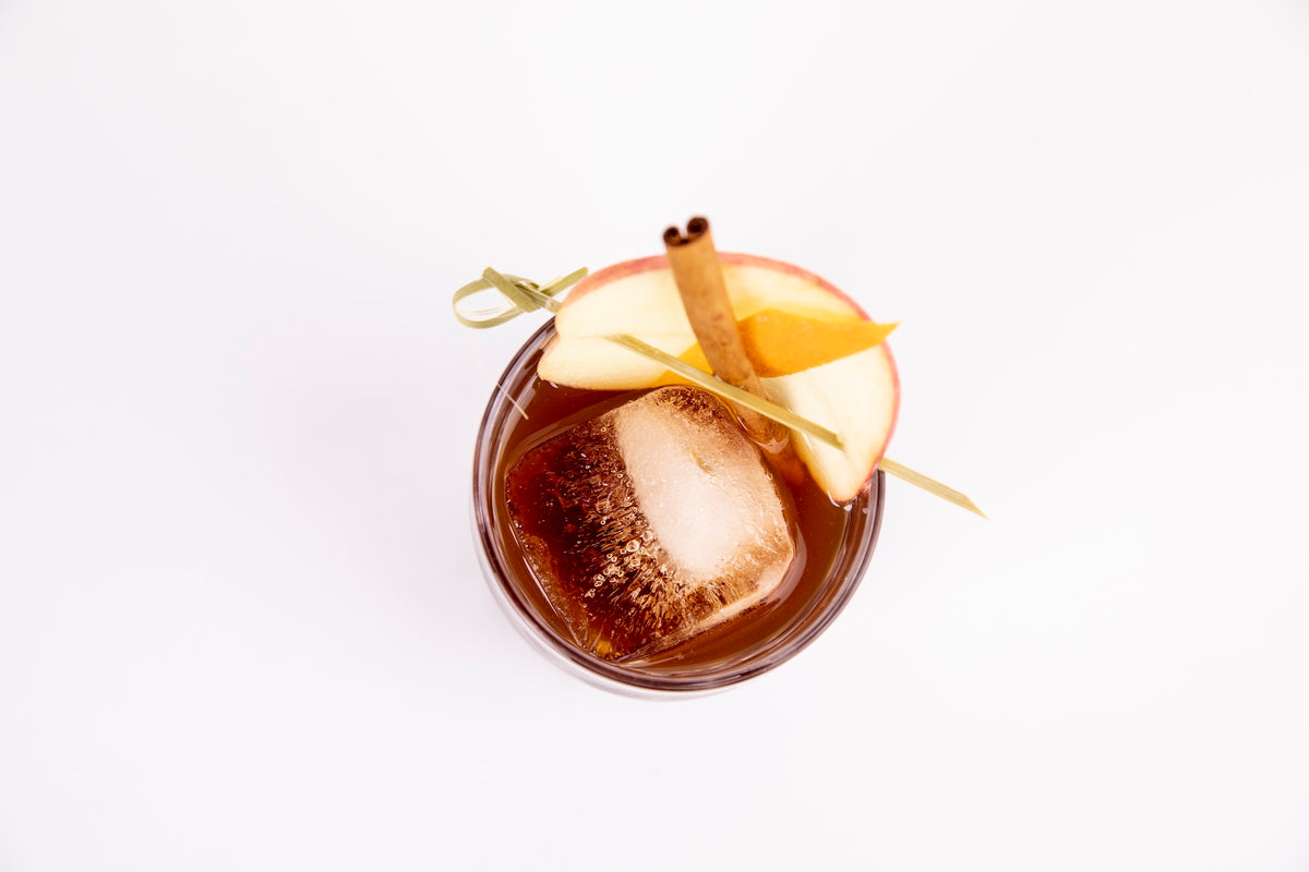 Cocktail Crate Spiced Old Fashioned cocktail over a large ice cube, garnished with apple, cinnamon stick and orange peel, seen from above on a white backdrop