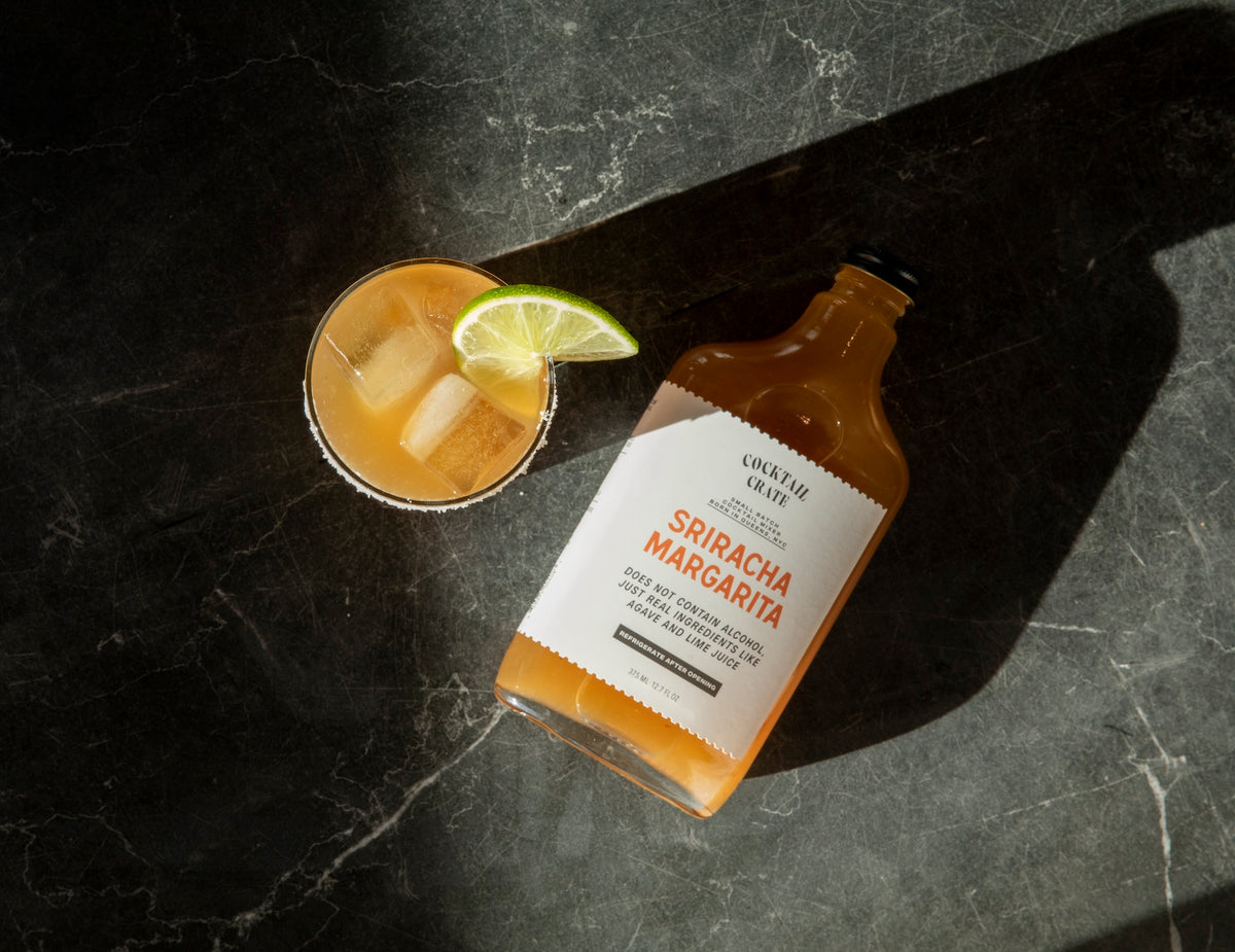 Cocktail Crate Sriracha Margarita mixer bottle beside a Sriracha Margarita cocktail with a lime wedge on a dark stone counter