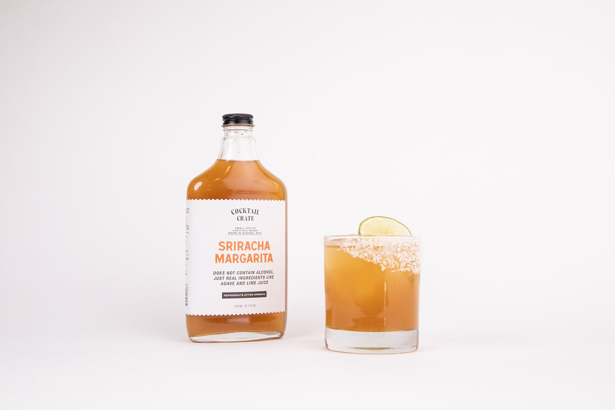 Cocktail Crate Sriracha Margarita mixer bottle beside a Sriracha Margarita cocktail with a lime slice on a white backdrop